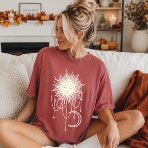 Könnte beinhalten: Ein altrosa T-Shirt mit kurzen Ärmeln und einem cremefarbenen Grafikdruck, der eine Sonne mit Gesicht mit geschlossenen Augen, hängenden Ketten und einer Mondsichel zeigt. Das T-Shirt hat einen Rundhalsausschnitt und eine lockere Passform.