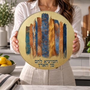May include: A round, gold-toned board featuring a vertical abstract brushstroke design in shades of blue, copper, and gold. The centre includes a floral pattern, and the Hebrew text "המוציא לחם מן הארץ" is printed in blue at the bottom.