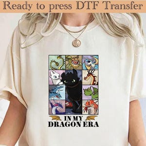 May include: Cream-colored t-shirt featuring a colorful dragon-themed graphic. The graphic includes various dragon illustrations in a grid pattern, with the text "IN MY DRAGON ERA" at the bottom. The top of the image reads "Ready to press DTF Transfer".