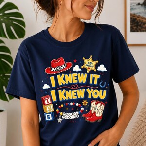 May include: A navy blue short-sleeve t-shirt featuring a graphic print with a red cowboy hat, a yellow sheriff badge with the text YEP, KNEW IT, red cowboy boots, alphabet blocks labeled T, S, 5, and a beaded bracelet with the text YOU & I FOREVER.