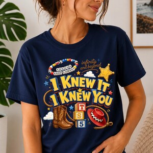 May include: A navy blue t-shirt featuring a graphic print with the text "I KNEW IT I KNEW YOU", "infinity and beyond", and "YOU'RE A GOOD COWBOY". The design includes a beaded bracelet, a gold star, a lasso, brown cowboy boots, and a red hat.