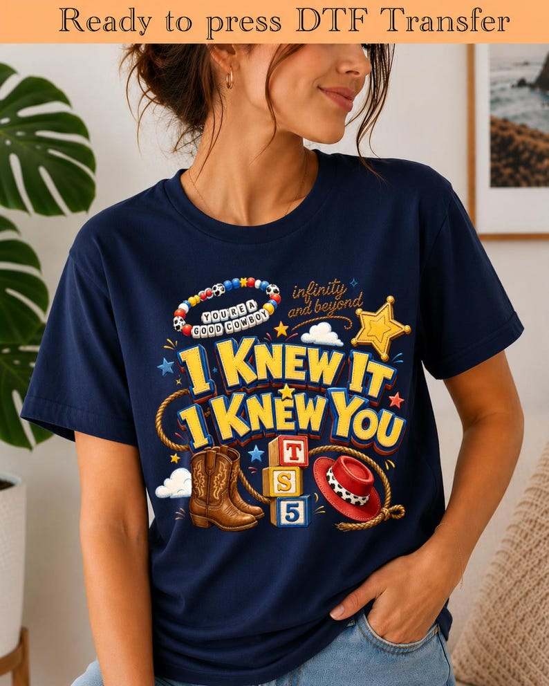 May include: A navy blue t-shirt featuring a graphic print with the text "I KNEW IT I KNEW YOU", "infinity and beyond", and "YOU'RE A GOOD COWBOY". The design includes a beaded bracelet, a gold star, a lasso, brown cowboy boots, and a red hat.