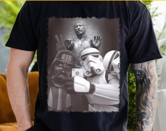 May include: A black t-shirt featuring a black and white image of three Star Wars characters taking a selfie. The characters are Darth Vader, a Stormtrooper, and another Stormtrooper. The image is set against a backdrop of a wall with a figure of Han Solo frozen in carbonite.