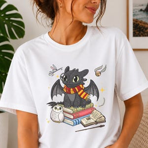 May include: A white t-shirt featuring a graphic of a black dragon wearing round glasses and a red and yellow striped scarf, sitting on a stack of books next to a white owl, a flying golden snitch, a key, an open book, and a wooden wand.