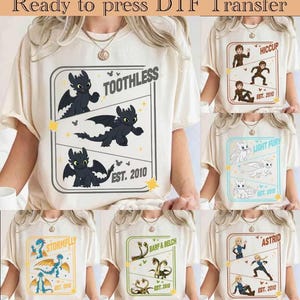 May include: Cream-colored t-shirts featuring various cartoon dragon designs. One shirt displays Toothless, another Hiccup, and others showcase Light Fury, Stormfly, Barf & Belch, and Astrid. Each design includes the text "EST. 2010". The phrase "Ready to press DTF Transfer" is at the top.