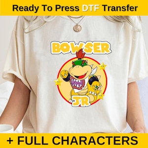 May include: A white t-shirt with a yellow circle containing a cartoon image of Bowser Jr. from the Super Mario Bros. video game series. The text "BOWSER JR" is printed in yellow letters above the image. The text "+ FULL CHARACTERS" is printed in yellow letters at the bottom of the image.