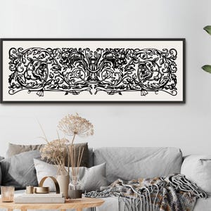 May include: A black and white framed artwork with an ornate, symmetrical design hangs above a gray sofa. The artwork features a detailed floral and scroll pattern. A large potted plant and a wooden coffee table complete the living room setting.