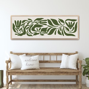 May include: A framed canvas art piece featuring a botanical design in shades of green on a cream background. The artwork is displayed above a wooden bench with pillows, and a potted plant. The bench has two pillows, one with the text "welcome home".