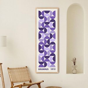 May include: A framed Bauhaus 1919 print features a geometric pattern in shades of purple. The artwork is displayed on a cream-colored wall, alongside a wooden chair with woven seat and a tripod floor lamp. A small, woven ottoman sits on the floor.