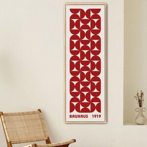 May include: A framed print with a red geometric pattern and the text "Bauhaus 1919". The print is hanging on a white wall in a room with a wooden floor, a wooden chair, and a floor lamp.