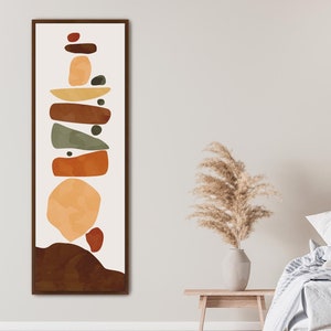 May include: Abstract art print featuring a stack of colorful geometric shapes in shades of brown, orange, green, and yellow. The print is framed in a brown wood frame and is hanging on a white wall.