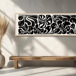 May include: A framed abstract art piece with a black and white floral design. The artwork features organic shapes and patterns, creating a modern aesthetic. The frame is a light wood color, and the artwork is displayed above a wooden bench.