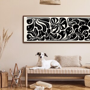 May include: A long, rectangular framed artwork with a black and white abstract design. The artwork features organic shapes and patterns. A dog is lying on a bench in front of the artwork.