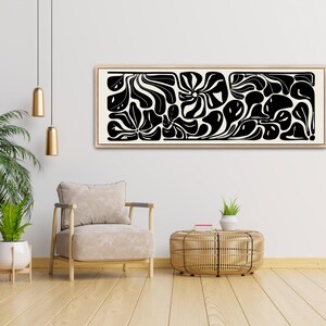 May include: A framed abstract art piece with a beige background and black organic shapes. The artwork is rectangular and hangs above a beige armchair and a woven side table. The room has a modern, minimalist aesthetic with wooden floors and a potted plant.
