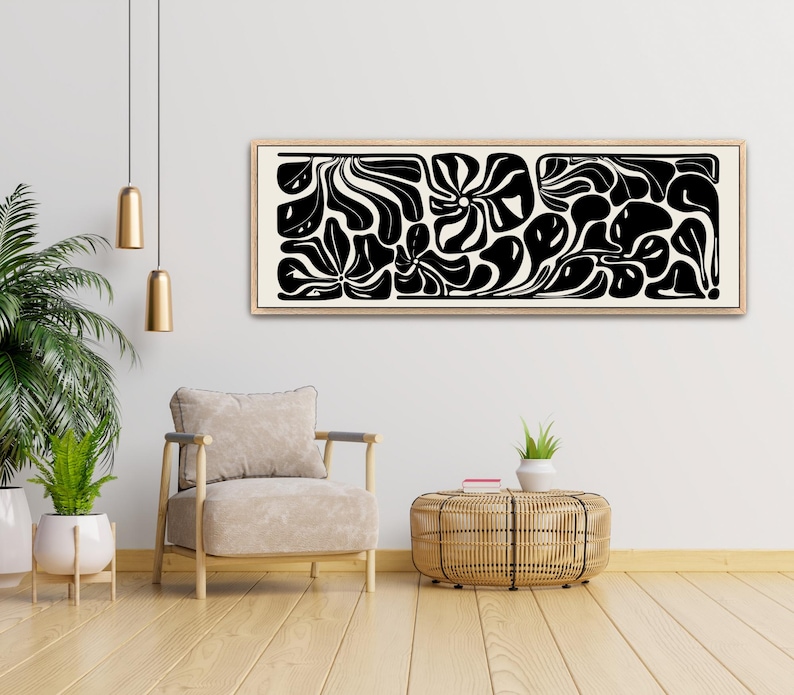 May include: A framed abstract art piece with a beige background and black organic shapes. The artwork is rectangular and hangs above a beige armchair and a woven side table. The room has a modern, minimalist aesthetic with wooden floors and a potted plant.