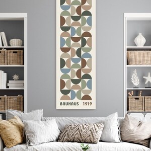 May include: A vertical wall art piece with a geometric design of semi-circles in shades of brown, green, and blue. The text "BAUHAUS 1919" is printed at the bottom. The artwork is displayed in a living room setting.