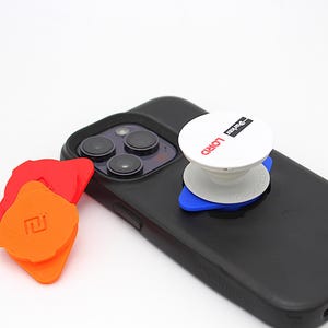 Puede incluir: Un teléfono inteligente negro con un soporte circular blanco adherido a la parte posterior, con el texto "Parker LORD" en negro y rojo. Junto al teléfono hay dos accesorios de agarre triangulares adicionales en colores naranja y rojo.