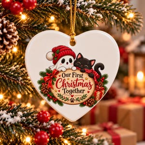 Può includere: Un ornamento in ceramica bianca a forma di cuore appeso a un cordino dorato. Raffigura un fantasma con un cappello rosso e un gatto nero con una sciarpa rossa, con il testo Our First Christmas Together, circondato da rami di pino e pigne.