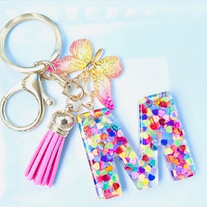 Puede incluir: Un llavero dorado con una borla rosa y la letra "M" de resina transparente llena de confeti en forma de corazón colorido. También incluye un dije de mariposa decorativo y un cierre.