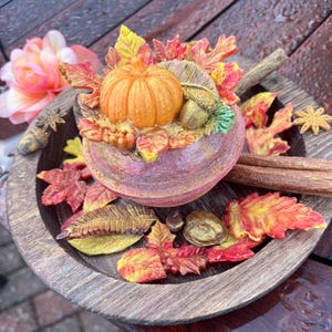 May include: A small, orange pumpkin surrounded by fall leaves and acorns in a brown pot with a glittery finish. The pot is sitting on a wooden tray with more fall leaves.