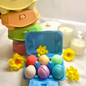 Könnte beinhalten: Ein Stapel bunter Eierkartons in Gelb, Orange, Grün und Rosa. Ein blauer Eierkarton enthält sechs bunte Badebomben. Gelbe Narzissen und brennende Kerzen sind ebenfalls zu sehen.