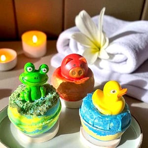 Könnte beinhalten: Drei bunte Badebomben mit Tieraufsätzen. Ein grüner Frosch sitzt auf einer grün-blauen Bombe, ein rotes Schwein auf einer braunen Bombe und eine gelbe Ente auf einer blau-gelben Bombe. Die Bomben stehen auf einem Teller mit brennenden Kerzen im Hintergrund.