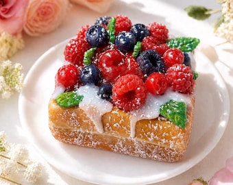 May include: A square pastry topped with fresh raspberries, blueberries, and red currants, drizzled with white icing and dusted with powdered sugar. The dessert sits on a white plate, with a floral background.