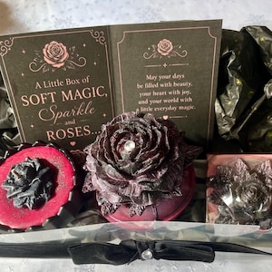 May include: A gift box with a black bow, containing a dark red rose-shaped bath bomb, a red soap with a rose design, and a small book with the text "A Little Box of SOFT MAGIC, Sparkle and ROSES..."