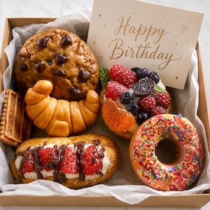 May include: A selection of baked treats in a gift box, including a chocolate chip cookie, croissant, and pastries. One pastry is topped with berries, another with raspberries and chocolate, and a donut with sprinkles. A card reads "Happy Birthday".