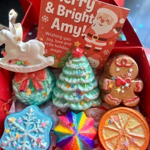 Könnte beinhalten: Eine festliche Geschenkbox mit weihnachtlichen Badebomben und Ornamenten. Enthält ein weißes Schaukelpferd, einen türkisfarbenen Weihnachtsbaum, einen Lebkuchenmann, einen Schneeflocke, einen Regenbogenstern und eine Orangenscheibe. Die Box enthält auch eine Karte mit "Merry & Bright Amy!"