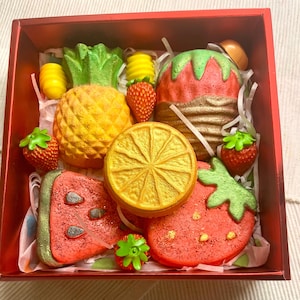 Könnte beinhalten: Eine rote Geschenkbox gefüllt mit bunten Badebomben in Form von Früchten. Die Badebomben sind Ananas, Orange, Wassermelone und Erdbeere.