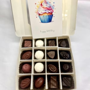 Peut inclure: Une boîte blanche avec une carte qui dit "joyeux anniversaire" et une illustration aquarelle d'un cupcake. La boîte est ouverte et contient 16 chocolats assortis dans une grille.