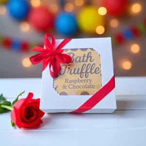 May include: A white gift box tied with a red ribbon, featuring a label that reads "Bath Truffle Raspberry & Chocolate." A red rose lies next to the box. The background has blurred colorful lights.