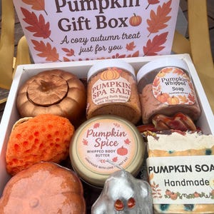 May include: A white box containing autumnal bath and body products. The box includes a pumpkin-shaped candle, bath salts, whipped soap, body butter, a sponge, and a bar of pumpkin soap. The box is labelled "Pumpkin Gift Box" with the text "A cozy autumn treat just for you."