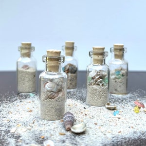 May include: Five small glass bottles with cork stoppers filled with sand and seashells. The bottles are arranged in a row on a gray surface with sand scattered around them.