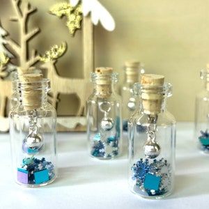 May include: Small glass bottles with cork stoppers, each containing decorative elements. The bottles feature silver bell charms and are filled with blue, purple, and silver confetti, including snowflake shapes and small cubes. A festive holiday theme.