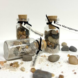 May include: Three small glass bottles with cork stoppers, filled with sand and pebbles. Each bottle has a tag that reads "Time Management". The bottles are tied with black ribbon and are surrounded by loose pebbles and sand.