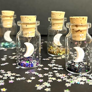 May include: Four small glass bottles with cork stoppers, each containing a white crescent moon charm and colorful glitter. The bottles are arranged on a black surface with scattered silver glitter stars.