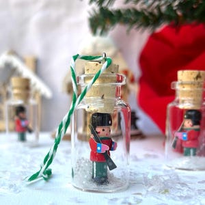 May include: A small glass bottle ornament with a cork top and a red and green toy soldier inside. The bottle is filled with white glitter and has a green and white twine tied around the top.