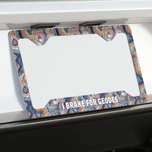 May include: A rectangular license plate frame featuring a colorful geode pattern in shades of blue, orange, and white. The bottom center of the frame displays the white text I BRAKE FOR GEODES in a clean, sans-serif font against the patterned background.