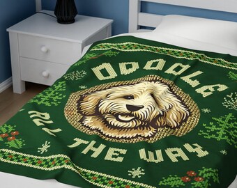 May include: A green blanket with the text "Doodle All The Way" and a graphic of a golden doodle dog. The blanket has a festive design with snowflakes, Christmas trees, and holly berries. The blanket is on a white bed.