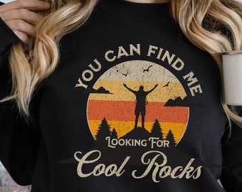 May include: Black sweatshirt with a vintage-style graphic of a person standing on a mountain with a sunset behind them. The text reads "You can find me looking for cool rocks."