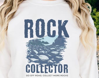 May include: White sweatshirt with a blue graphic that reads "ROCK COLLECTOR." The graphic depicts a collection of rocks and the text "GO OFF ROAD, COLLECT MORE ROCKS" below the word "COLLECTOR."