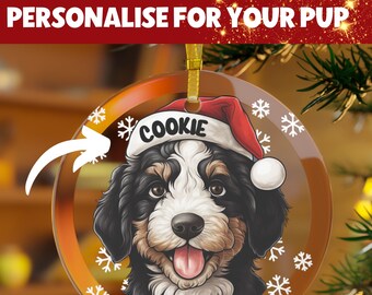 May include: A round glass Christmas ornament with a white background and snowflakes. A cartoon image of a Bernedoodle dog wearing a Santa hat is in the center of the ornament. The dog's name, "Cookie", is written on the hat.