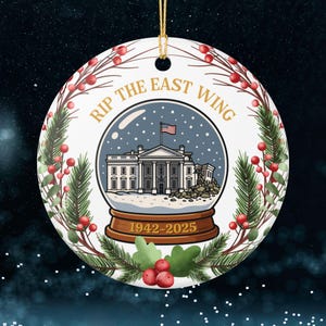 May include: A round, white ceramic ornament with a gold string for hanging. The ornament features a snow globe design with the White House and the text "RIP THE EAST WING 1942-2025". The ornament is decorated with a wreath of green pine needles and red berries.