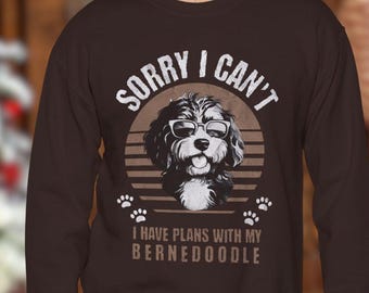 Può includere: Felpa marrone con la scritta "SORRY I CAN'T" sopra un'illustrazione in bianco e nero di un cane con gli occhiali. Il testo "I HAVE PLANS WITH MY BERNEDOODLE" è sotto.