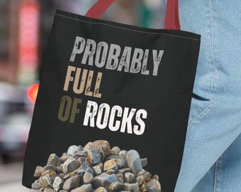 May include: Black tote bag with the text "PROBABLY FULL OF ROCKS" in white and tan lettering. The bottom of the bag features a pile of various rocks. The bag has red handles and is being held by a person wearing blue jeans.