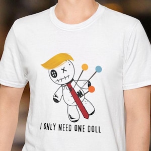 May include: White t-shirt featuring a cartoon voodoo doll with blonde hair, a red tie, and pins. The doll has a stitched mouth and an eye with a button. The text "I ONLY NEED ONE DOLL" is printed below the graphic.