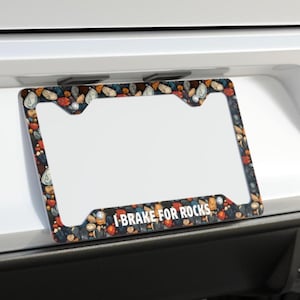 May include: License plate frame with a colorful pebble design and the text "I BRAKE FOR ROCKS" in white. The frame is rectangular with a white center and a border filled with various colored stones.