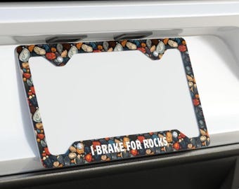 May include: Rectangular license plate frame featuring a pebble pattern border and the white text "I BRAKE FOR ROCKS". The frame has a white central area and is decorated with various coloured stones.